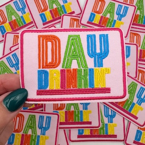 Pu&ograve; includere: Toppa ricamata rosa con le parole "DAY DRINKIN" in lettere colorate e in grassetto. La toppa ha un bordo rosa ed &egrave; tenuta da una mano con smalto verde acqua. Altre toppe simili sono sullo sfondo.