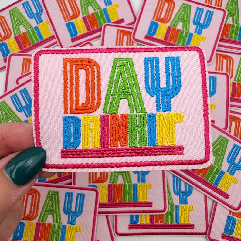 Pu&ograve; includere: Toppa ricamata rosa con le parole "DAY DRINKIN" in lettere colorate e in grassetto. La toppa ha un bordo rosa ed &egrave; tenuta da una mano con smalto verde acqua. Altre toppe simili sono sullo sfondo.