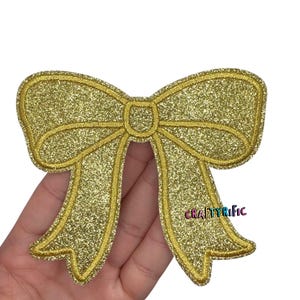 May include: A gold glitter bow applique with a stitched outline. The bow is held in a hand against a white background. The text "CRAFTYrific" is visible on the right side of the bow.