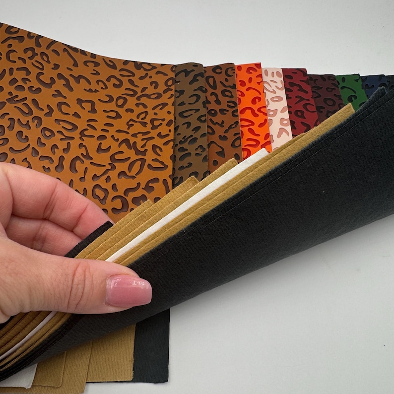 K&ouml;nnte beinhalten: Ein Stapel Kunstlederb&ouml;gen in verschiedenen Farben und Mustern, darunter Leopardenmuster, Orange, Rot, Braun und Schwarz. Die B&ouml;gen sind in der Mitte gefaltet und &uuml;bereinander gestapelt.