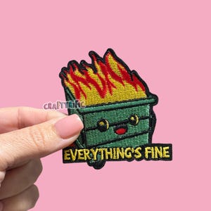 May include: An embroidered patch featuring a green dumpster with a smiling face, engulfed in bright yellow and red flames. Below the dumpster, a black rectangular banner contains the yellow embroidered text EVERYTHING'S FINE.