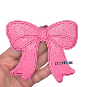 May include: A pink glittery bow-shaped patch with embroidered details. The patch has a classic bow design with two loops and two long tails. The text "CRAFTYRIFIC" is visible on the lower right side of the patch.