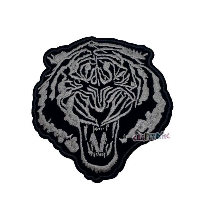 May include: Embroidered patch featuring a roaring tiger's head. The design showcases a detailed tiger face with open mouth and visible teeth, in shades of gray and black. The patch has a black border and the text "CRAFTYrific".