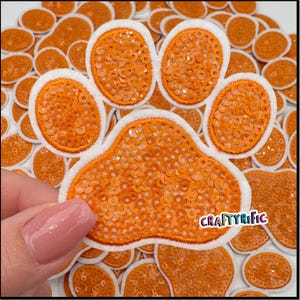 May include: Orange paw print iron-on patch with white stitching and sequins. The patch is surrounded by other paw print patches.