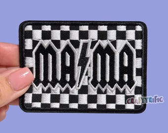 May include: A rectangular patch with a black and white checkered background. The patch features the word "MAMA" in bold, stylized black letters with a lightning bolt in the center. The patch is held by a hand.