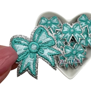May include: A close-up of a teal and silver glitter bow applique. The bow is embroidered with a light blue thread. The bow is in a heart-shaped container with other bows.