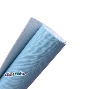 May include: A roll of light blue textured faux leather fabric with a crosshatch pattern. The fabric is partially unrolled, revealing a white backing. The logo "CRAFTYRIFIC" is visible in the lower left corner.