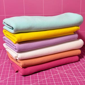 K&ouml;nnte beinhalten: Ein Stapel von sechs Waffelstrick-Stoffproben in verschiedenen Farben, darunter hellblau, gelb, Lavendel, wei&szlig;, Pfirsich und rosa. Der Stoff ist gefaltet und auf einer rosa Schneidematte gestapelt.
