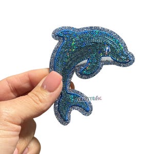 May include: A blue sequined dolphin patch with a silver glitter border. The dolphin has a detailed design with a white eye and is held by a hand. The text "Craftyrific" is visible.
