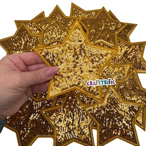 May include: Gold sequin star patches. The patches are star-shaped with a gold border and filled with gold sequins. The patches are being held by a hand. The text "CRAFTYRIFIC" is visible on one of the patches.