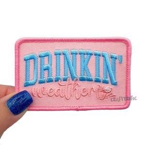 May include: A rectangular pink embroidered patch with the words "DRINKIN'" in blue block letters above "weather" in cursive. A small cup graphic is on the bottom right. The patch has a pink border.