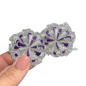 May include: Two embroidered cheerleading pom-pom patches. The patches feature a silver glitter border, white and purple accents, and a radial design. The patches are held by a hand. The text "CRAFTYRIFIC" is visible on the right patch.