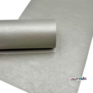 May include: A roll of silver-gray textured faux leather fabric. The fabric is partially rolled, revealing the underside. The word "CRAFTYrific" is printed in the lower right corner.