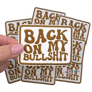 May include: White and brown embroidered patch with the text "BACK ON MY BULLSHIT". The patch has a rectangular shape with rounded corners and a brown border. Several patches are visible.