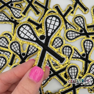 May include: Black and white lacrosse stick patches with gold glitter borders. The patches feature a detailed net design and are ideal for crafting projects. The image shows a hand holding one patch, with many more in the background.