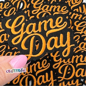 May include: A close-up of multiple black felt patches with orange embroidered text that reads "Game Day".