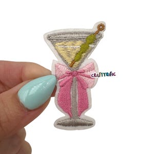 May include: Embroidered iron-on patch of a martini glass with a pink bow. The glass is filled with a yellow liquid and has a green olive on a toothpick. The text "CRAFTYRIFIC" is visible in the background.