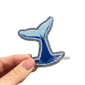 May include: A blue and silver glitter embroidered patch in the shape of a whale tail. The tail is a gradient of light to dark blue, with a silver glitter outline. The patch is held by a hand.