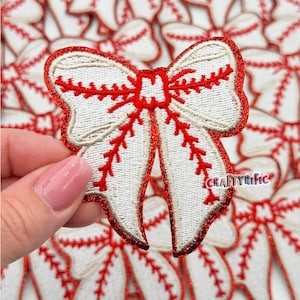 May include: White and red embroidered baseball-themed bow with a glittery finish. The bow is shaped like a baseball with red stitching.