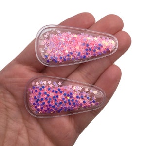 May include: Two clear plastic hair clips filled with pink and purple iridescent star-shaped glitter.
