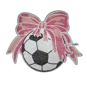 May include: A white and black soccer ball with a pink sequined bow on top. The bow is made of pink sequins and has a large, centered bow.