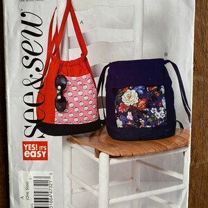 May include: A sewing pattern for two drawstring bags. The pattern is for one size and includes instructions for making a red bag with a pink and white cat print and a blue bag with a floral print. The pattern is from the See & Sew brand and is labeled "Yes! It's Easy".