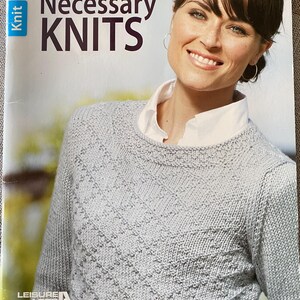 May include: A book cover titled "Necessary Knits" features a woman wearing a light gray knit sweater. The cover includes text: "Pullovers, cardigans, and vests you'll love!" and "by Susan Robicheau". The Leisure Arts logo is visible.