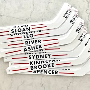 May include: A stack of white hockey stick shaped wooden signs with red and blue stripes and personalised names. The names include: Tavi, Sloan, Charlotte, Leo, River, Asher, Sydney, Kingston, Brooke, and Spencer. The signs also have the text "Westys Besties 2024" printed on them.