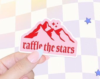 May include: A pink sticker featuring a mountain range design in red, with the text "raffle the stars" in a gothic font. The sticker is held by a hand with a light blue and white manicure. The background is a checkered pattern with blue star shapes.