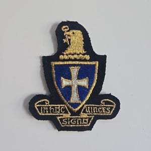 May include: Embroidered patch featuring a blue shield with a white cross, framed by gold trim and text. A gold eagle sits atop the shield. The patch is on a black background.