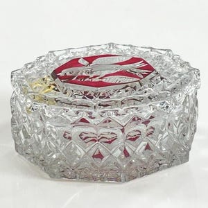 May include: A clear, faceted glass trinket box with a red interior and a red and white bird design on the lid. The box has an octagonal shape and a decorative, textured surface. The lid is also faceted.