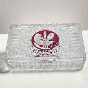 May include: A rectangular clear glass trinket box featuring a faceted diamond-cut pattern across its surface. Centered on the lid is a circular inset medallion displaying a deep magenta background with a white etched illustration of a bird.