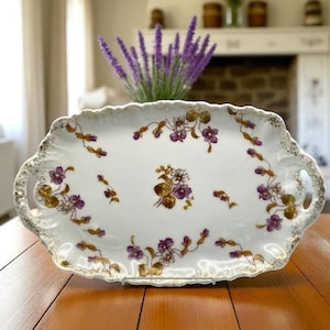 May include: An antique white porcelain serving tray with a scalloped edge and gold trim. The tray is decorated with purple and gold floral patterns. It has two handles and is placed on a wooden table.