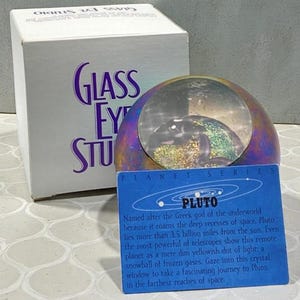 May include: A Glass Eye Studio Pluto planet series paperweight. The paperweight is a clear glass orb with a colorful interior, set on a metallic base. A blue card with text about Pluto is in front of the paperweight. A white box with the words "Glass Eye Studio" is on the left.