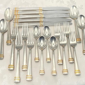 May include: A collection of silver-colored flatware with gold-colored accents. The set includes spoons, forks, and knives. The handles have a gold band near the base. The flatware is arranged on a white surface.