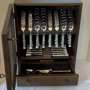 May include: A wooden cutlery cabinet with an open door, revealing a set of silverware. The silverware has a bamboo-style design. The cabinet has a drawer at the bottom with more silverware. The cabinet is brown.