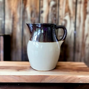 May include: A brown and white glazed ceramic pitcher with a handle. The pitcher is on a wooden surface.