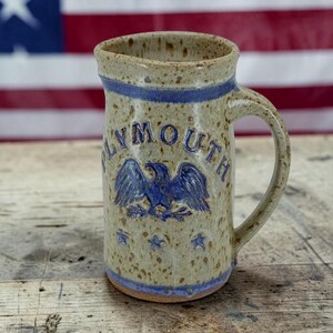 May include: A speckled beige ceramic pitcher with a blue rim and base. The front features the embossed blue text PLYMOUTH arched over a blue eagle emblem, with three blue stars below the eagle. It has a single handle on the right side.