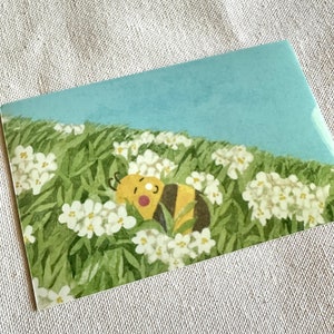 Op de afbeelding: Een ansichtkaart met een illustratie van een bij die rust in een veld met witte bloemen en groen gras onder een blauwe lucht. De bij heeft gele en zwarte strepen. De ansichtkaart is rechthoekig.