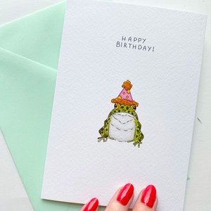 Może przedstawiać: Biała kartka urodzinowa z napisem "HAPPY BIRTHAY!" i akwarelową ilustracją zielonej żaby w czapce urodzinowej. Karta jest połączona z jasnozieloną kopertą, idealną do wysyłania życzeń urodzinowych.