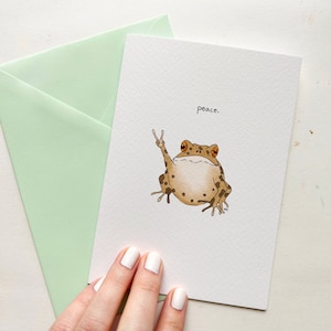 Könnte beinhalten: Eine Grußkarte mit einem braunen und weißen Frosch, der das Friedenszeichen zeigt. Auf der Karte steht das Wort "peace".