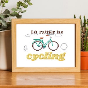 May include: A framed cross-stitch artwork with the text "I'd rather be cycling" and a bicycle illustration. The artwork is in a wooden frame and is displayed on a wooden surface with plants and a camera.