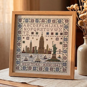 Peut inclure: Une broderie encadrée représentant l'horizon de New York, avec la Statue de la Liberté et des voiliers. Le motif est dans des tons de marron, vert et bleu, avec le texte "NEW YORK - ANNO 1842" en bas. Le cadre est en bois clair.