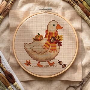 Op de afbeelding: Een kruissteekproject met een gans met een sjaal en herfstbladeren en een pompoen. De borduursel zit in een houten borduurring, omringd door borduurgaren in verschillende kleuren, een schaar en een patroonvel. Het kleurenpalet omvat beige, bruin, oranje en groen.