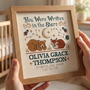Puede incluir: Un bordado de punto de cruz enmarcado con el texto "You Were Written in the Stars" y el nombre "Olivia Grace Thompson". El diseño incluye un oso, un zorro, una luna y estrellas. También se incluye la fecha y el peso de nacimiento. El marco es de madera clara.