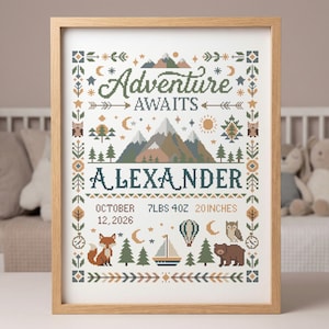 May include: A framed cross-stitch art piece with the text "Adventure Awaits" and the name "Alexander". The design includes mountains, trees, and woodland creatures. The frame is light brown. The artwork measures 20 inches.