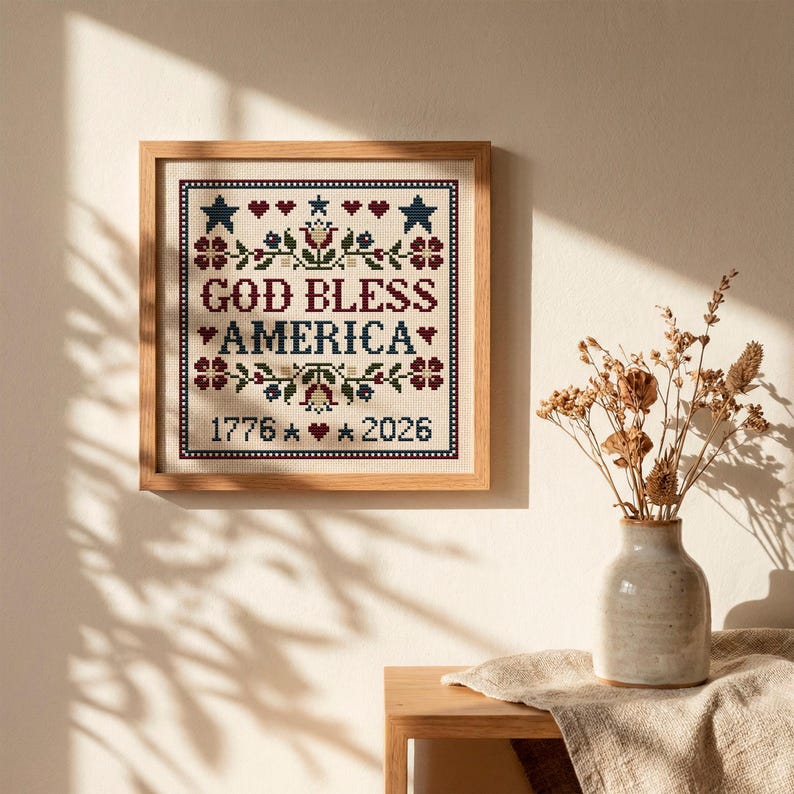 May include: A framed cross-stitch with the text "GOD BLESS AMERICA", and the dates 1776 and 2026. The design includes stars, hearts, and floral motifs in red, blue, and white. The frame is made of light-coloured wood.