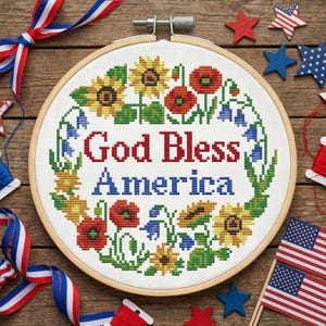 Op de afbeelding: Een borduurring met kruissteek met de woorden "God Bless America" in rood en blauw. Het ontwerp bevat zonnebloemen, klaprozen en andere bloemen. Patriotische sterren, linten en miniatuur Amerikaanse vlaggen omringen de ring.