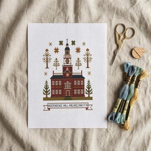 May include: A cross-stitch of Independence Hall in Philadelphia, Pennsylvania, with a banner reading "INDEPENDENCE HALL-PHILADELPHIA 1776." The image includes embroidery floss, scissors, and a needle threader.
