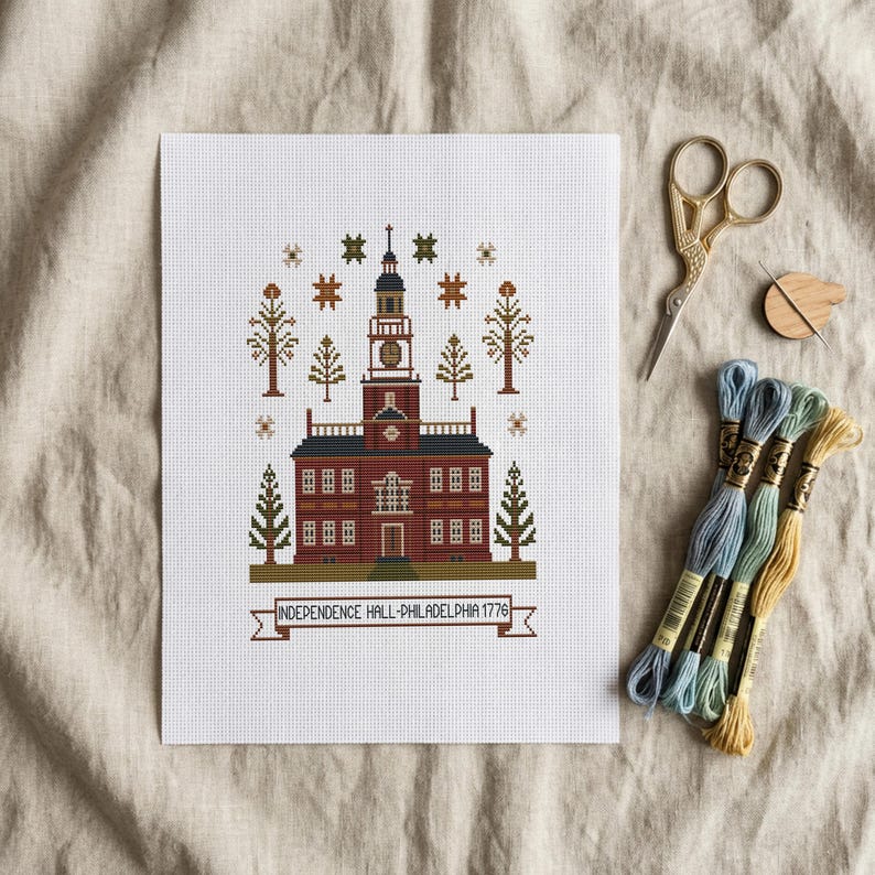 May include: A cross-stitch of Independence Hall in Philadelphia, Pennsylvania, with a banner reading "INDEPENDENCE HALL-PHILADELPHIA 1776." The image includes embroidery floss, scissors, and a needle threader.